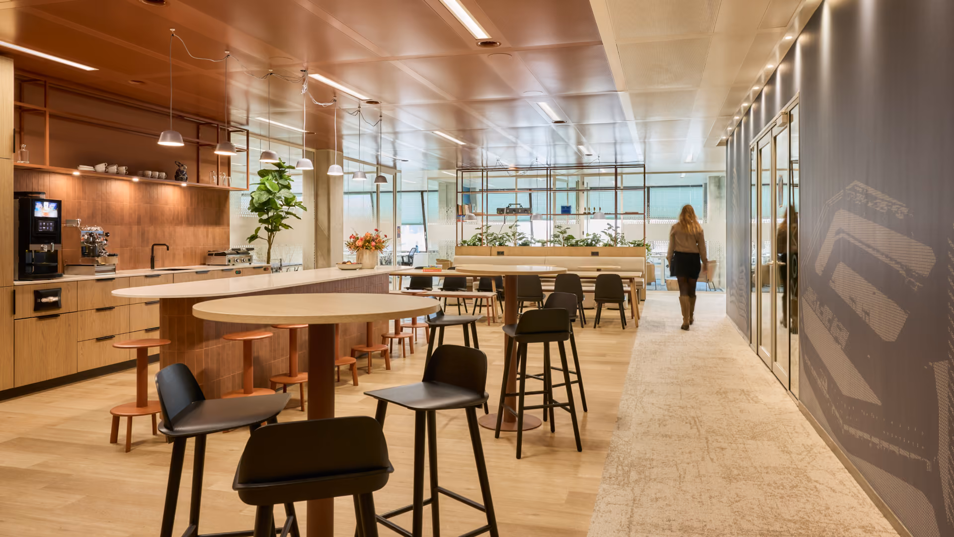 Modern kantoorruimte met houten vloeren en meubels, ronde en rechthoekige tafels met hoge stoelen, planten en een koffieautomaat aan de linkerkant, een vrouw loopt aan het einde van de gang.
