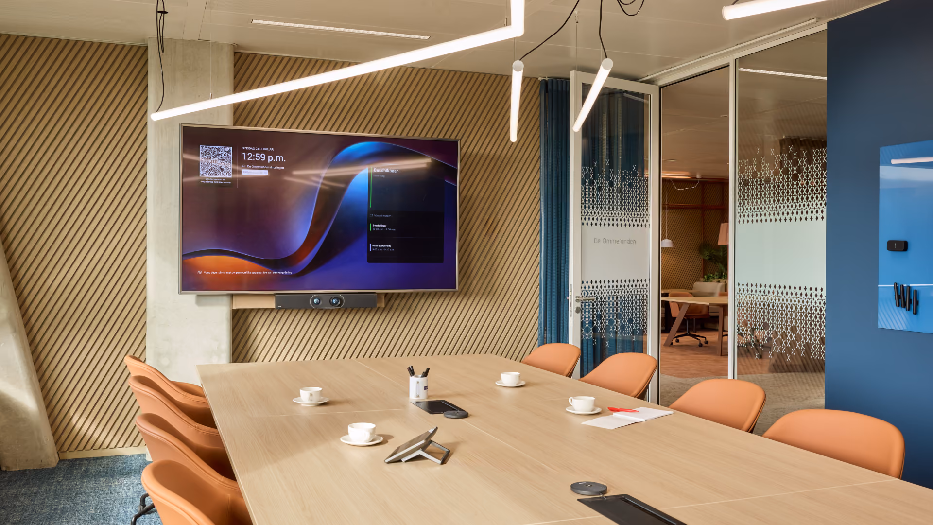 Moderne vergaderruimte met een houten tafel omringd door acht oranje stoelen, een grote wandmonitor aan een houten panelenwand toont een scherm met tijd 12:59 p.m., en minimalistische verlichting hangt aan het plafond.