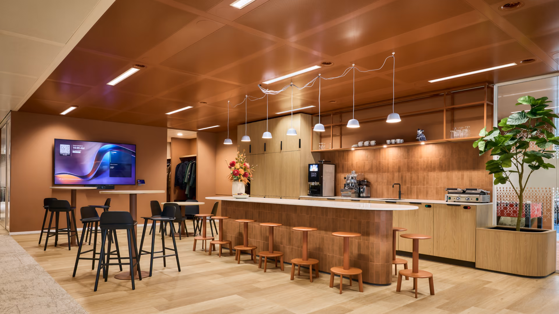 Moderne kantoorkeuken met lange bar en houten krukken, hoge tafels met zwarte barkrukken, inbouwapparatuur, planten en een tv-scherm aan de muur.