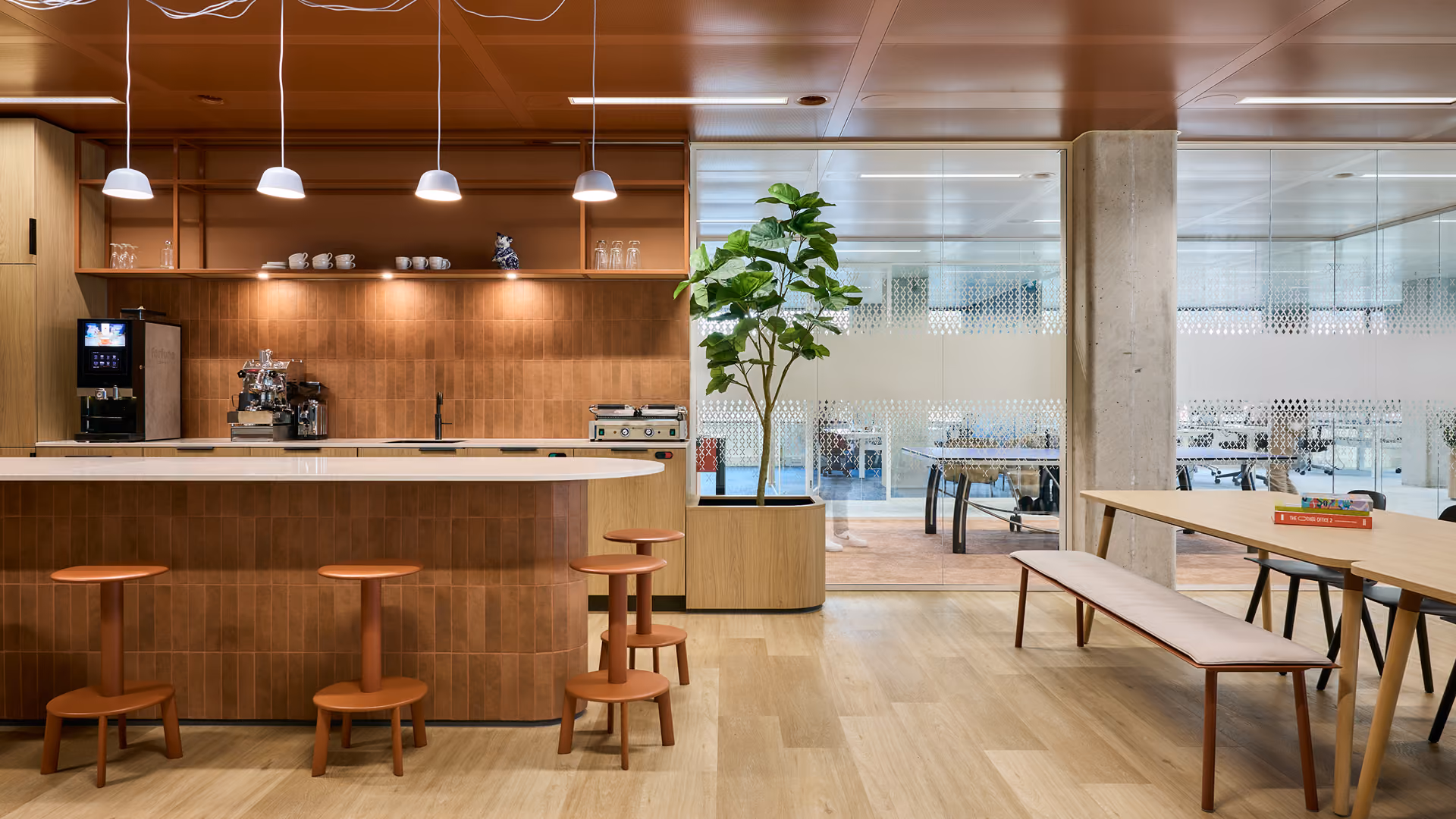 Moderne kantoorruimte met houten bar en krukken, koffiemachine, kantoorplanten en lichte vergadertafel met banken.