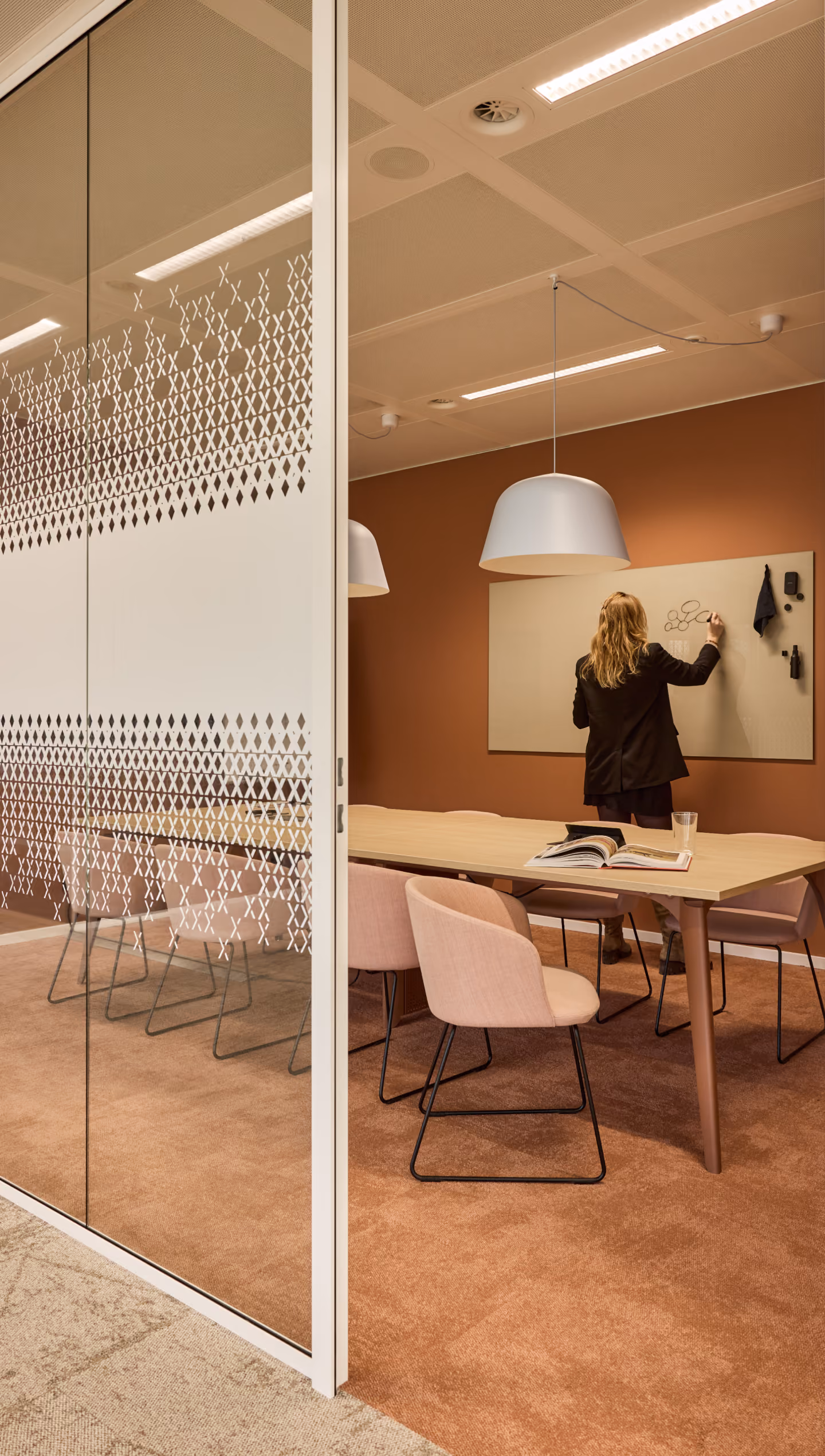 Kantoorruimte met een glazen wand met patroon aan de linkerkant, een houten tafel met roze stoelen en een vrouw die op een beige krijtbord aan de bruine muur tekent.