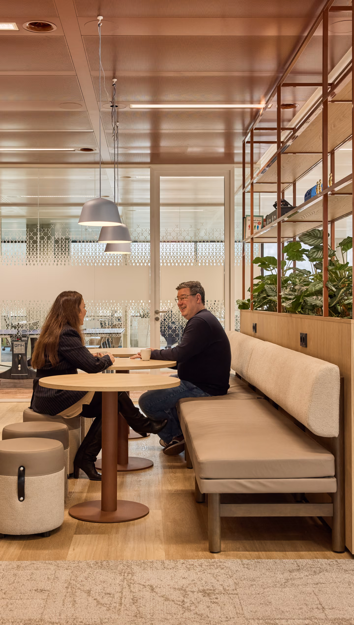 Twee personen zitten aan een ronde tafel in een moderne, lichte kantoorruimte met houten vloer, banken en planten op een open rek naast hen.