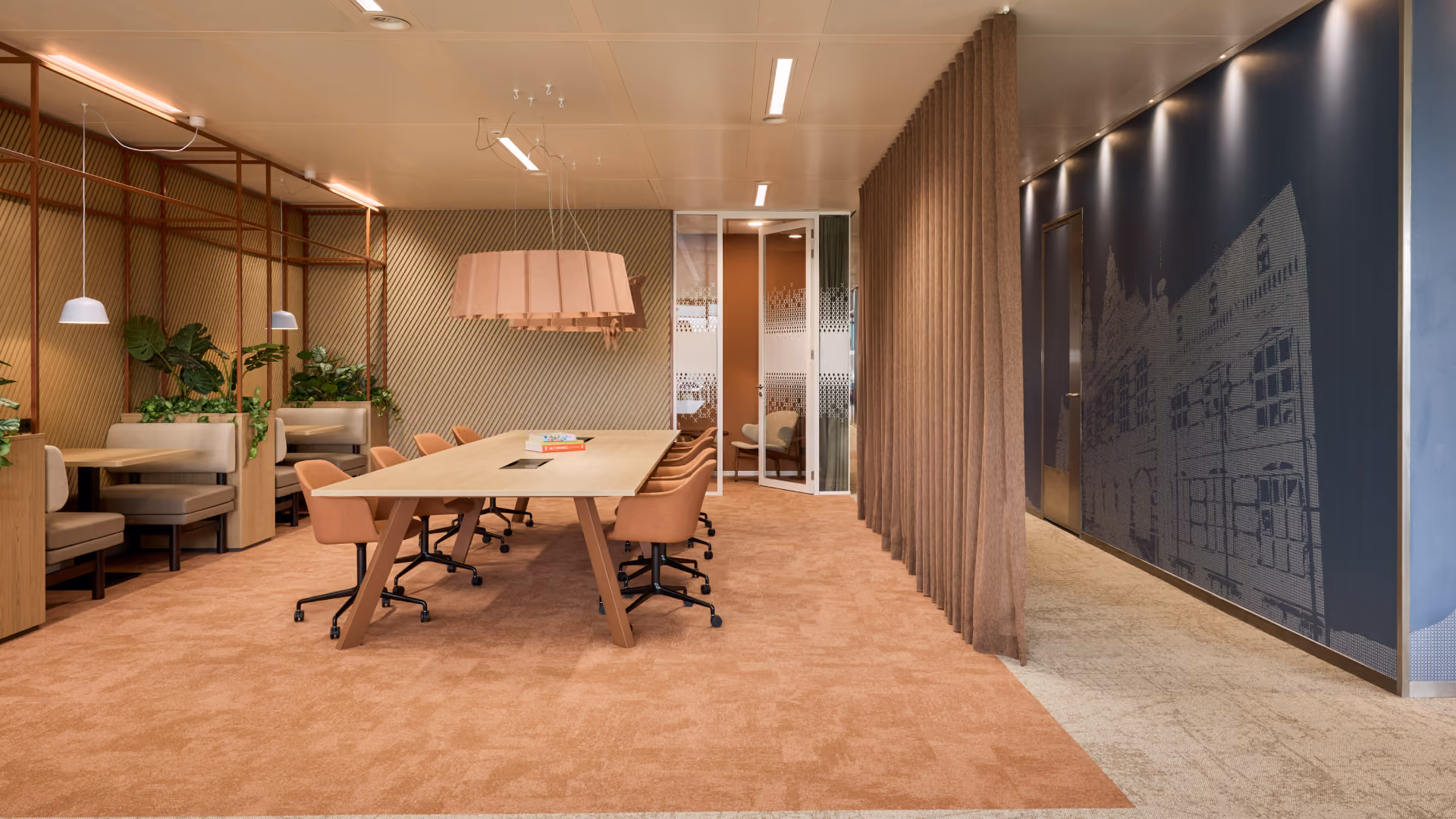 Moderne vergaderruimte met lange houten tafel, roze bureaustoelen, ingebouwde banken en planten aan de linkerkant, zachte verlichting en een glazen deur aan de achterzijde.