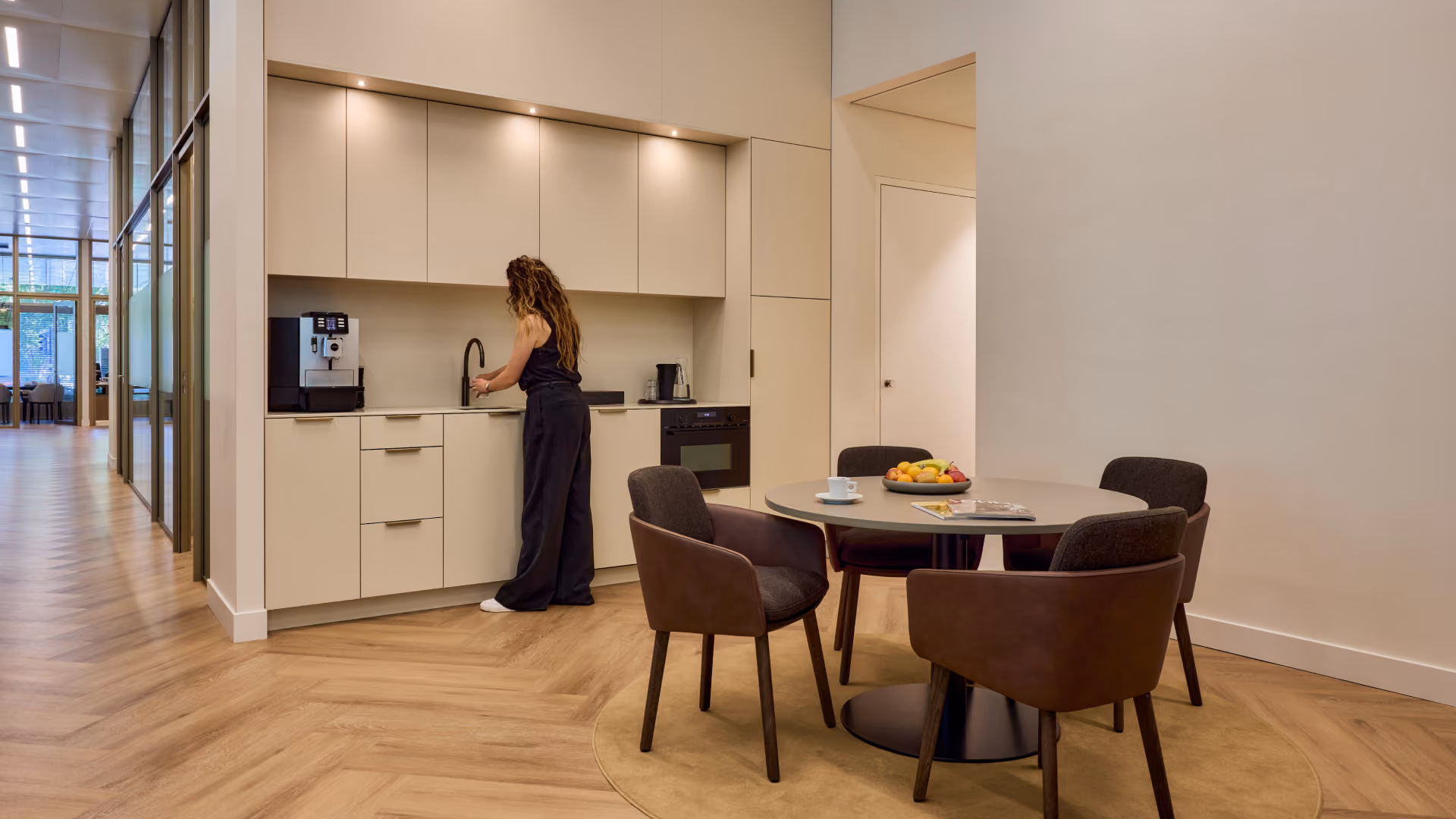 Een vrouw staat in een moderne kantoorkeuken met lichte kasten en een koffiezetapparaat, terwijl ze water bij de gootsteen tapt naast een ronde tafel met vier stoelen en een fruitschaal in een lichte, ruime kamer.