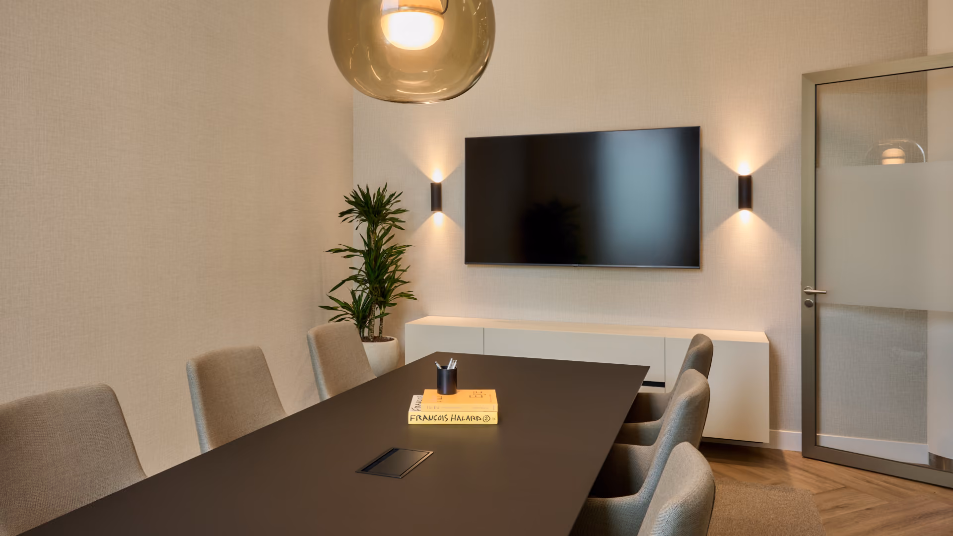 Moderne vergaderruimte met een lange zwarte tafel, zes grijze stoelen, een boek en pennenhouder op tafel, een grote plantenpot, een wandkast, een flatscreen-tv en sfeerverlichting aan de muur.