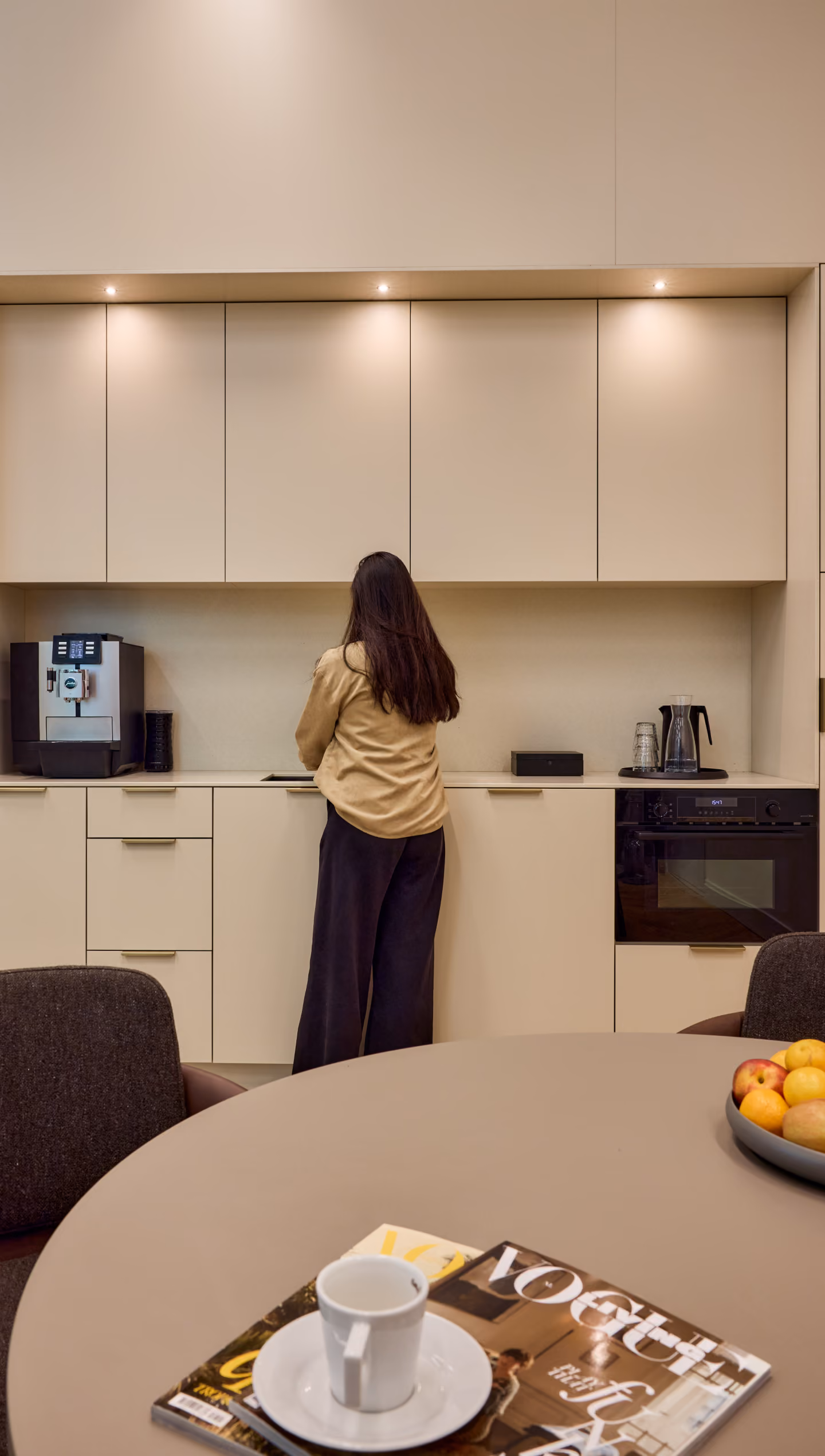 Vrouw staat met rug naar de camera in een moderne keuken, omringd door beige kasten, een koffiemachine links, een oven rechts en een tafel met een tijdschrift en een kopje koffie op de voorgrond.