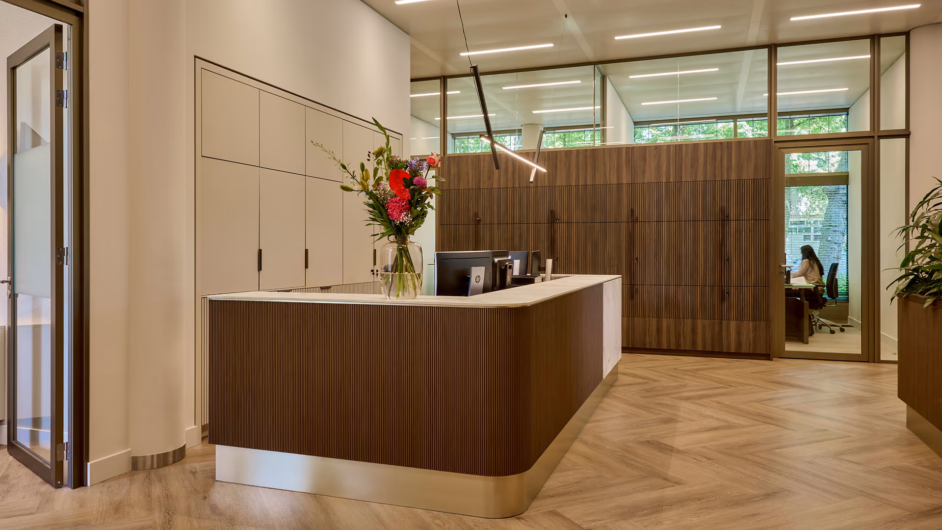 Moderne receptie met een houten balie, een vaas met bloemen, ingebouwde kasten en glazen wanden waarachter iemand aan een bureau zit.