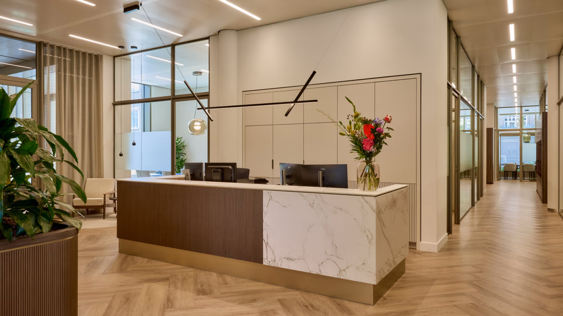 Modern receptiebalie in een kantoor met marmer en houtafwerking, twee computerschermen, een vaas met kleurrijke bloemen, grote groene planten, glazen wanden, en een lange gang met veel natuurlijk licht.