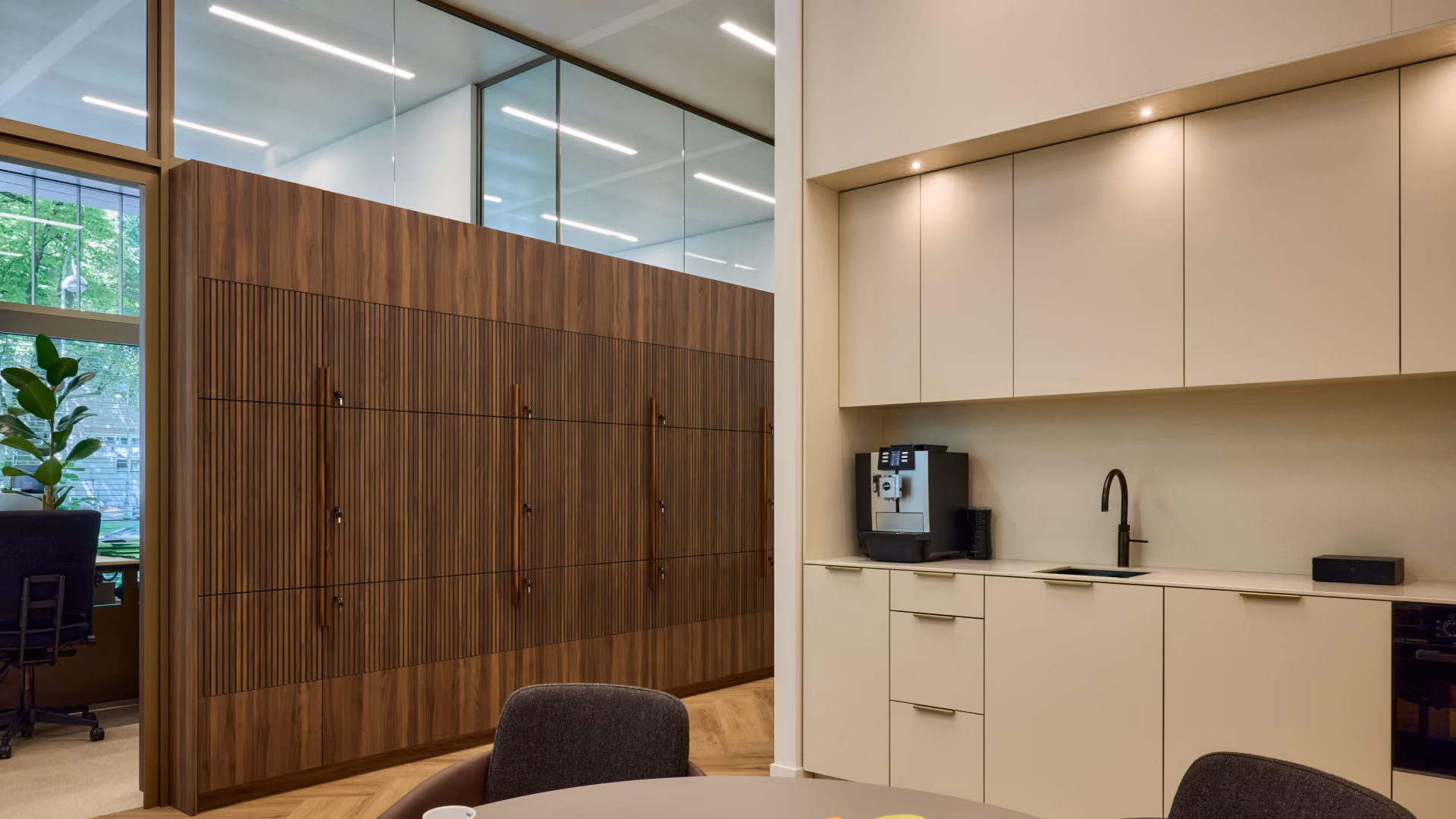 Moderne kantoorruimte met een ronde tafel en stoelen op de voorgrond, een koffiezetapparaat en wastafel in een lichtkleurige kitchenette, en glazen wanden met houten panelen aan de linkerkant.