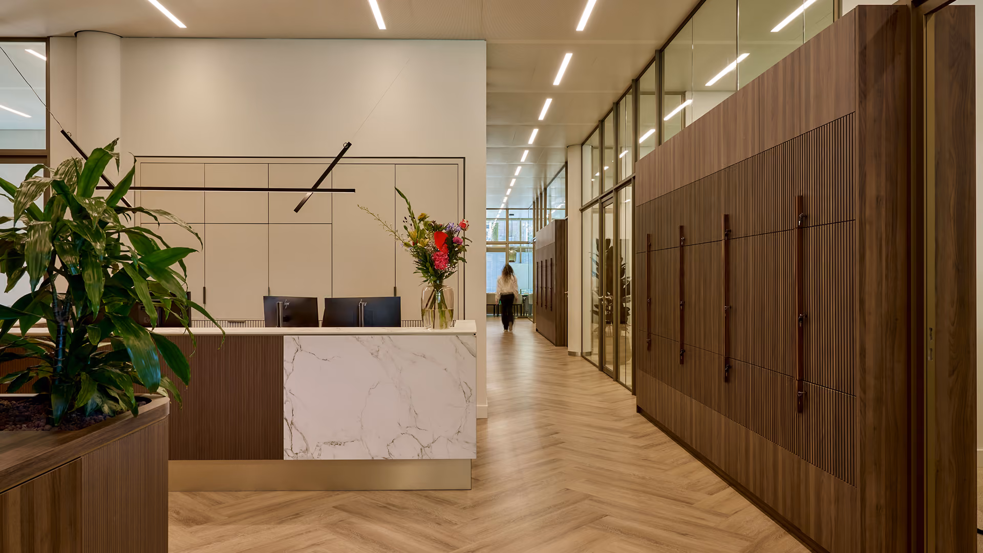 Moderne kantoorruimte met houten vloer in visgraatmotief, witte receptiebalie met marmerlook, planten en bloemstuk, glazen wanden en houten panelen aan de rechterkant.
