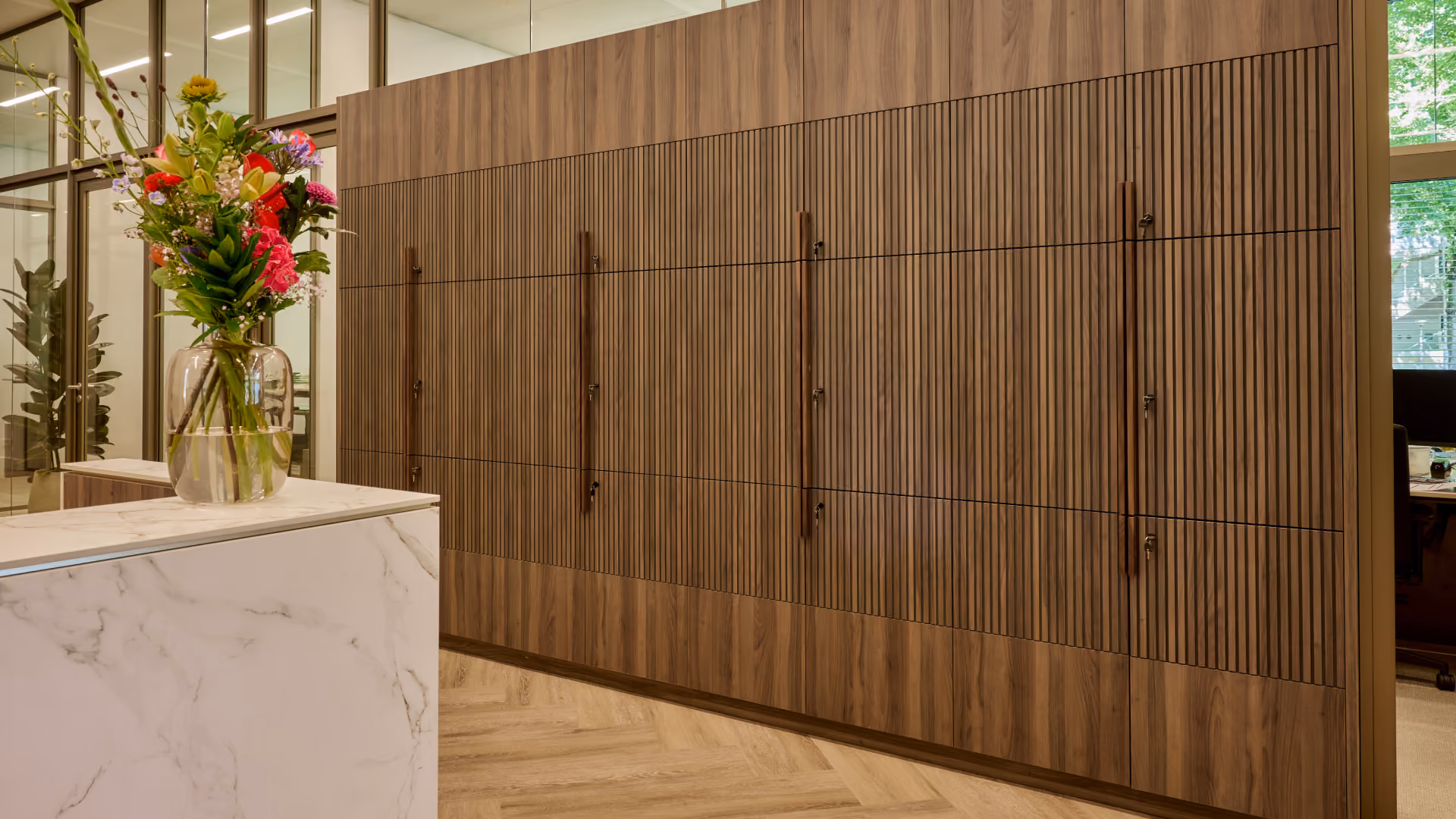 Moderne kantoorruimte met marmeren balie en een grote vaas met kleurrijke bloemen, daarnaast een wand van houten kasten met verticale lamellen en metalen handgrepen.