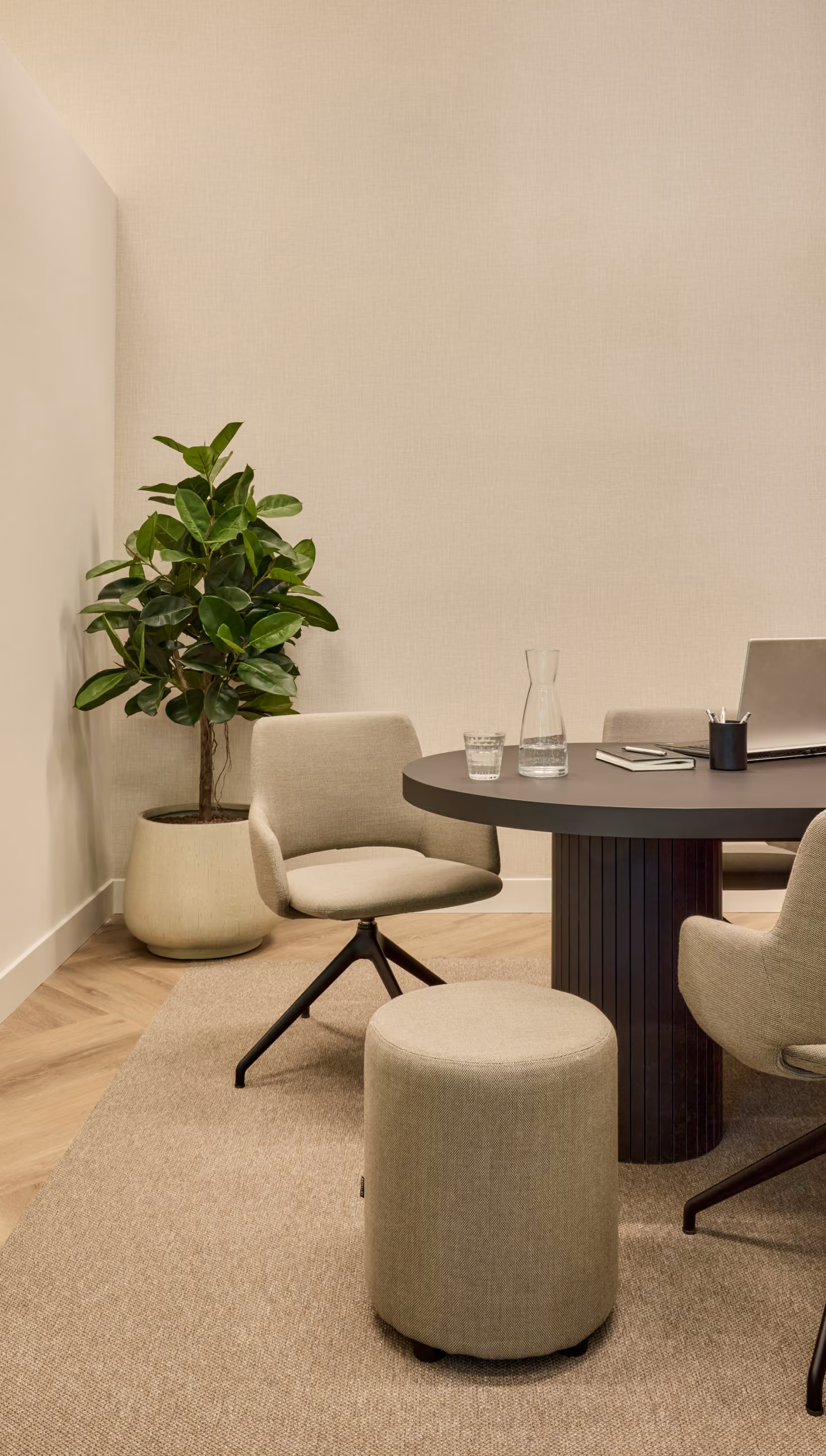 Moderne vergaderruimte met een ronde zwarte tafel, beige gestoffeerde stoelen en een kruk, een glazen karaf en glas water, een laptop en kantoorbenodigdheden op de tafel, en een grote groene kamerplant in een pot in de hoek.