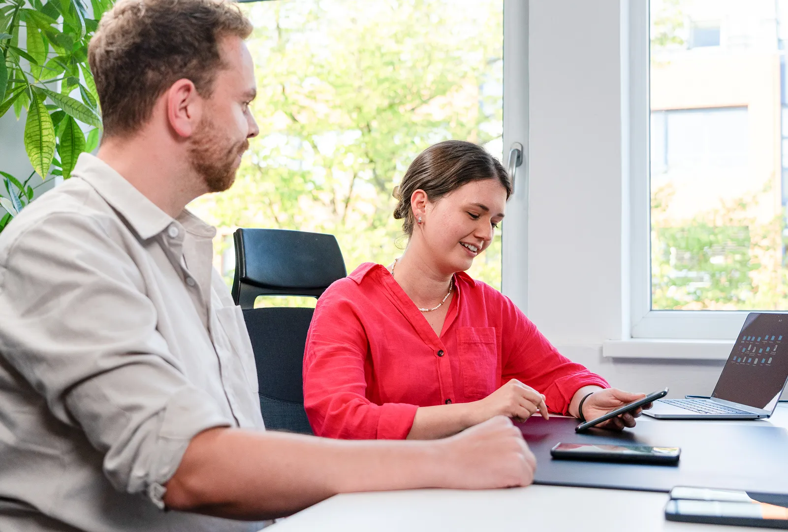 Zwei Personen sitzen an einem Tisch und testen Software, eine Frau in rotem Hemd nutzt ein Tablet, ein Mann blickt zu ihr.