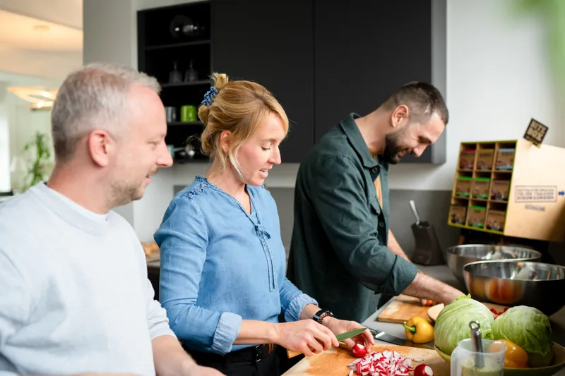 Drei Personen in einer Küche schneiden Gemüse, eine Frau schneidet Radieschen, ein Mann schneidet andere Gemüsesorten.