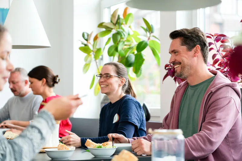 Menschen sitzen lächelnd zusammen an einem Tisch und essen in einem hellen Raum mit Pflanzen.