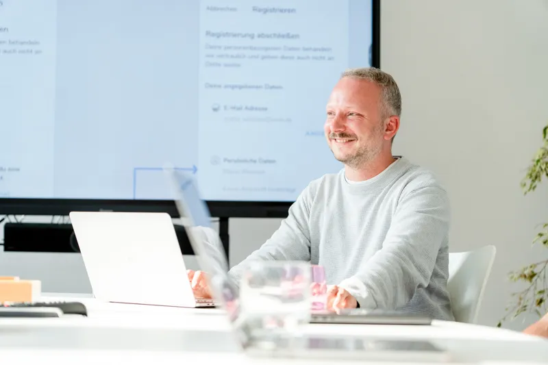 Lachender Mann sitzt an einem Tisch vor einem Laptop mit einem Bildschirm im Hintergrund, der ein App-Design anzeigt.