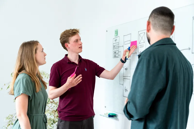 Drei Personen diskutieren vor einem Whiteboard mit Skizzen und Haftnotizen in einem hellen Büro.