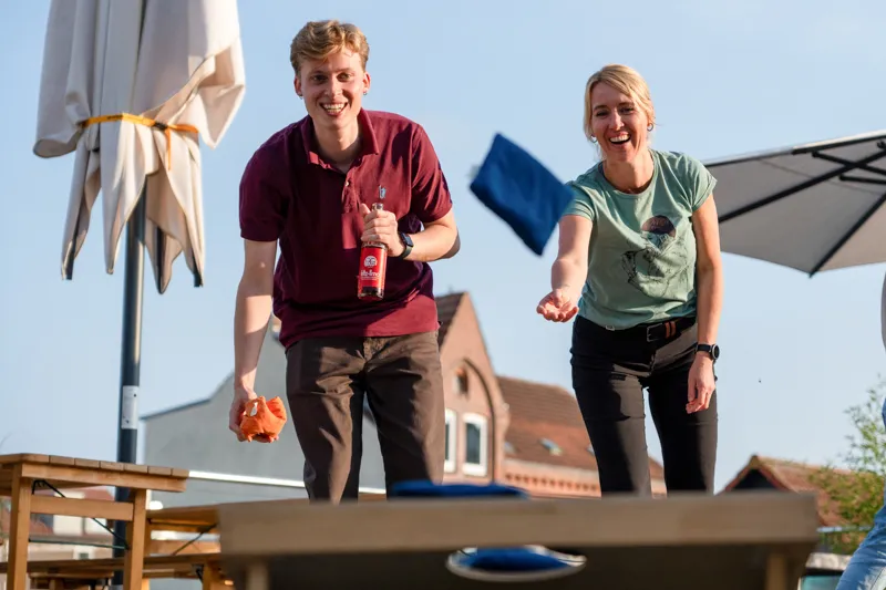 Zwei lachende Personen spielen Cornhole im Freien, eine hält eine Flasche und die andere wirft ein blaues Säckchen.