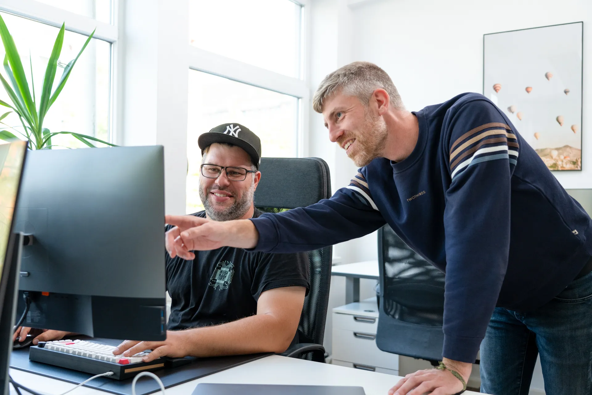 Zwei Männer arbeiten gemeinsam an einem Computer im hellen Büro, einer sitzt und der andere zeigt auf den Bildschirm.