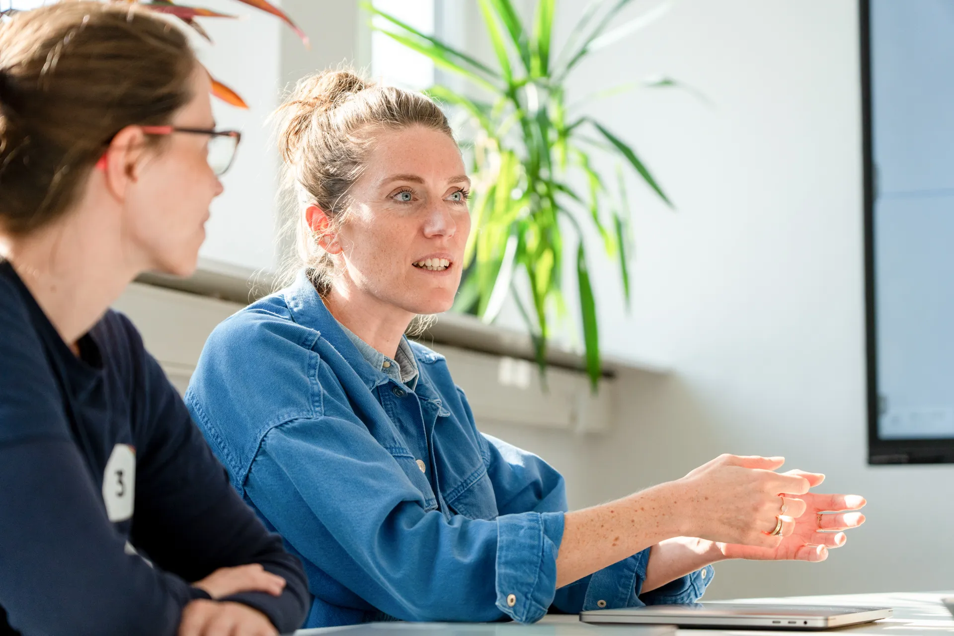 Zwei Frauen sitzen an einem Tisch, eine spricht und gestikuliert, im Hintergrund eine grüne Pflanze.