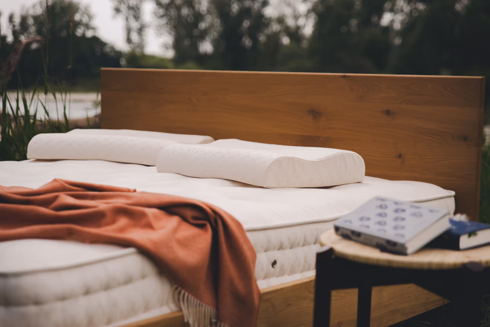 Letto con materasso e cuscini Materia, con una coperta rossa appoggiata e di fianco uno sgabello con dei libri, foto in esterna