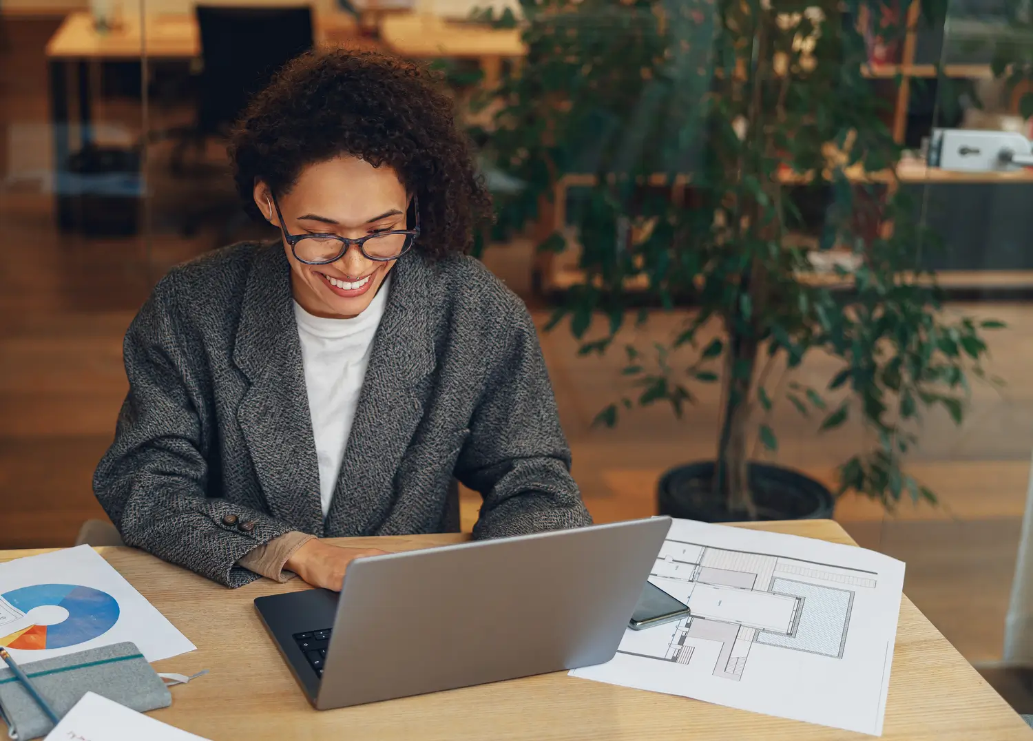 An adult woman with curly hair and glasses smiles as she works on a laptop at her desk in an office.