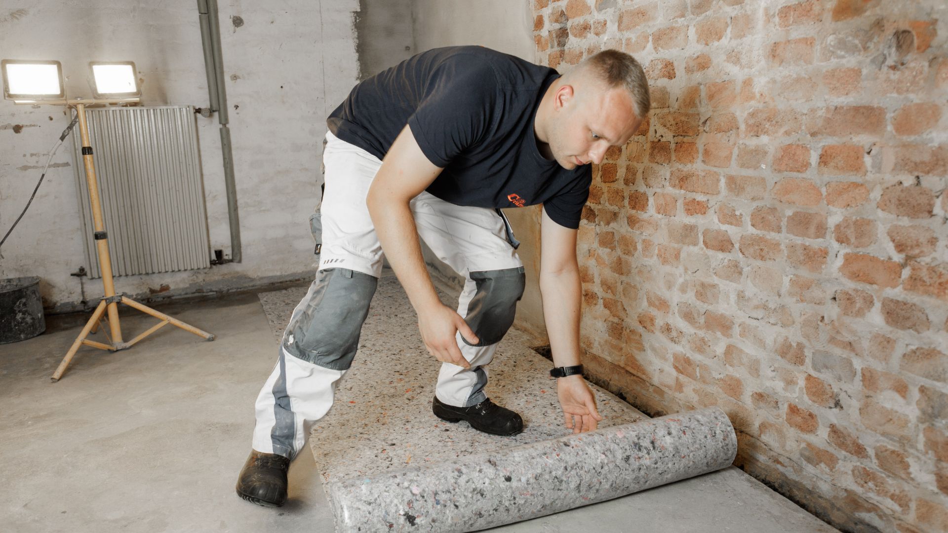 Handwerker rollt Schutzmatte auf Kellerboden vor Ziegelwand aus.