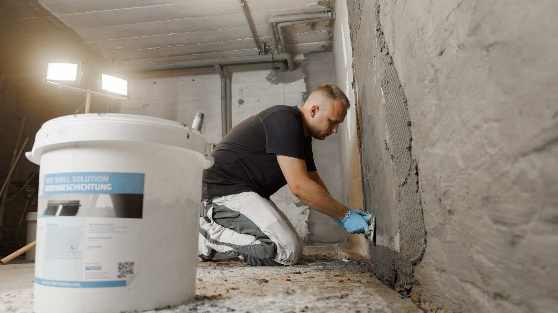 Ein Handwerker trägt in einem Keller nach Wasserschäden eine Abdichtungsbeschichtung auf die Wand auf, um Feuchtigkeit nach Starkregen zu beseitigen.