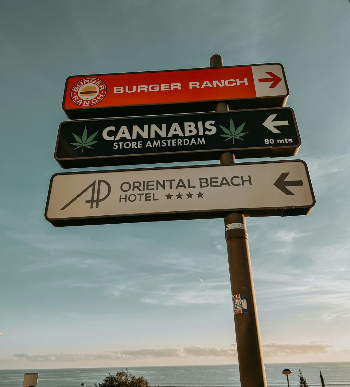 Signpost displaying directions to a burger range, the Amsterdam Store Cannabis, and the Oriental Beach Hotel in Porto, Portugal.