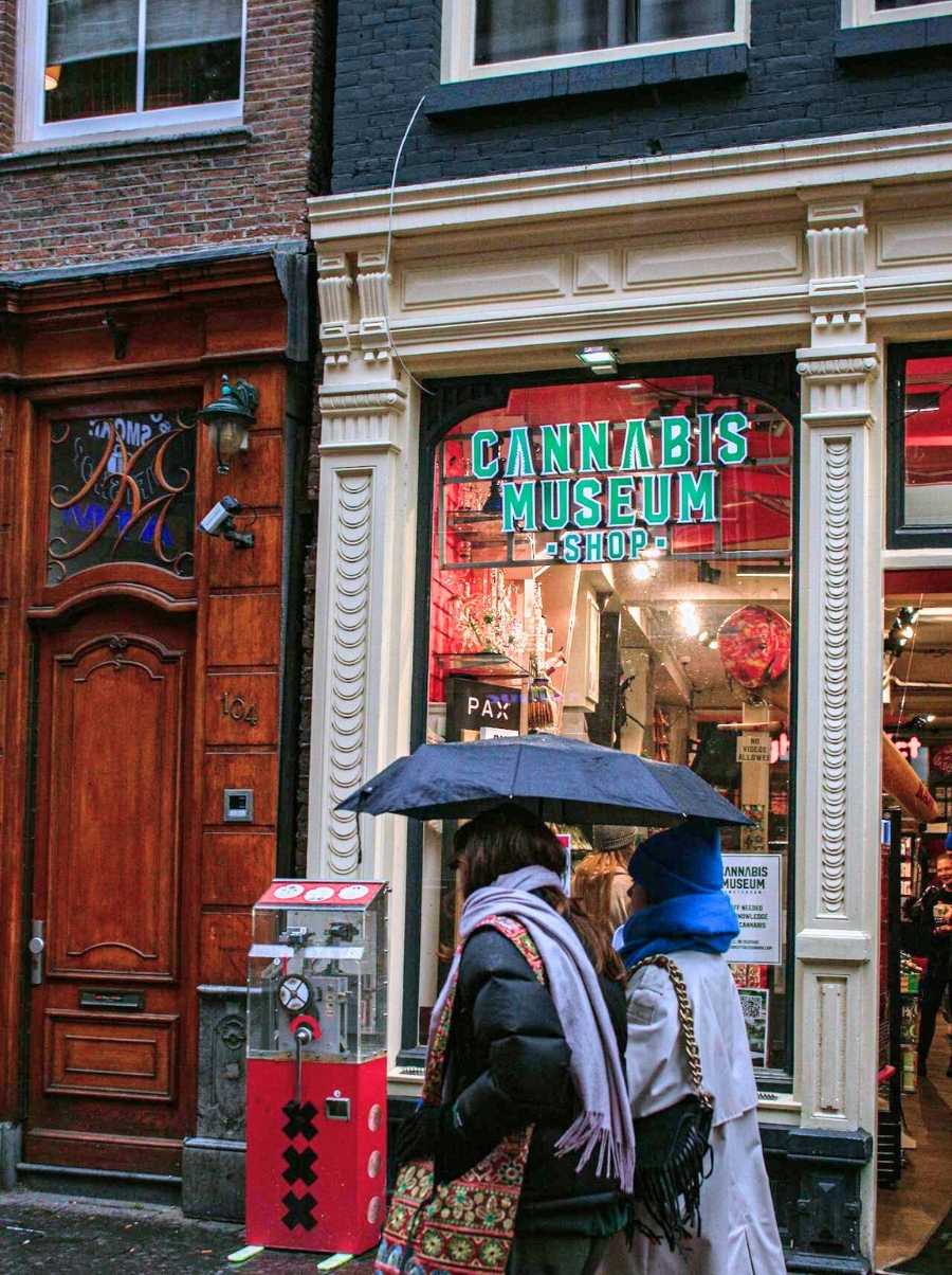 Outside of a cannabis store in Amsterdam, with two women walking past, one of them holding an umbrella.
