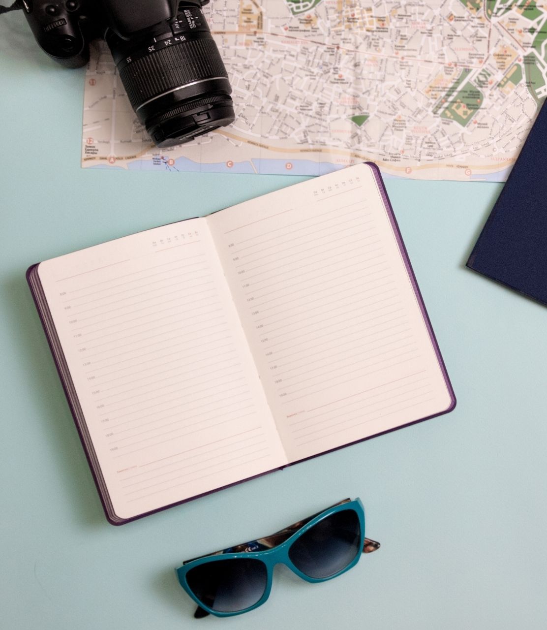 Flat lay of travel items arranged on a blue background, displaying sunglasses, an open notebook, a camera, and a map.