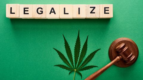 Overhead view of a cannabis leaf and a wooden gavel resting on a green background, with the word "LEGALIZE" spelt out above the leaf using individual wooden letter blocks