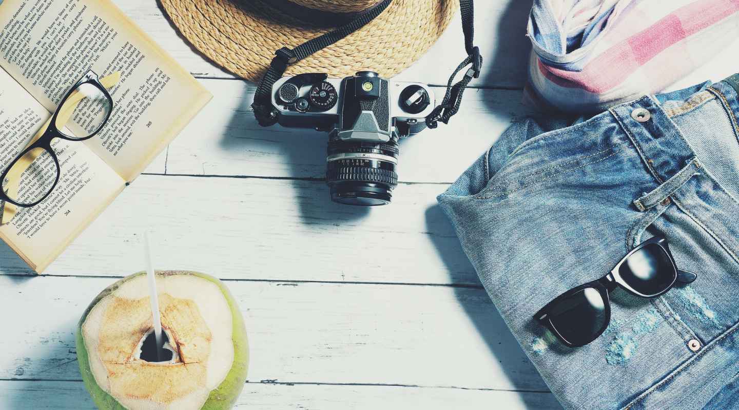 Flatlay of assorted travel items, including an open coconut, a book, sunglasses, a camera, and a pair of jeans on a white wooden surface