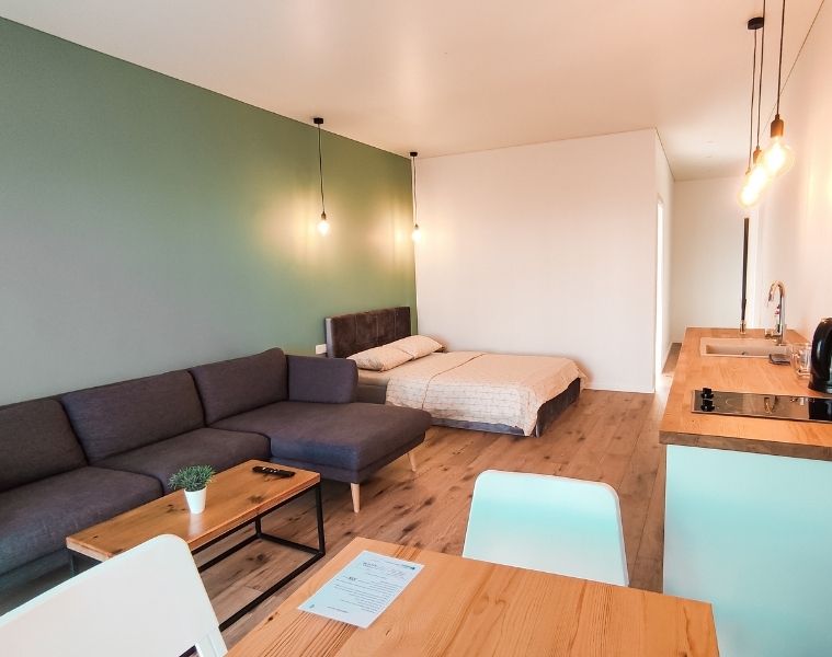 Interior of a small studio flat with a double bed, L-shaped sofa, table, and kitchen area inside.