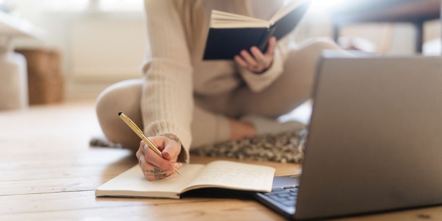 A woman sitting in front of a laptop, writing in a notepad while holding a book in her other hand