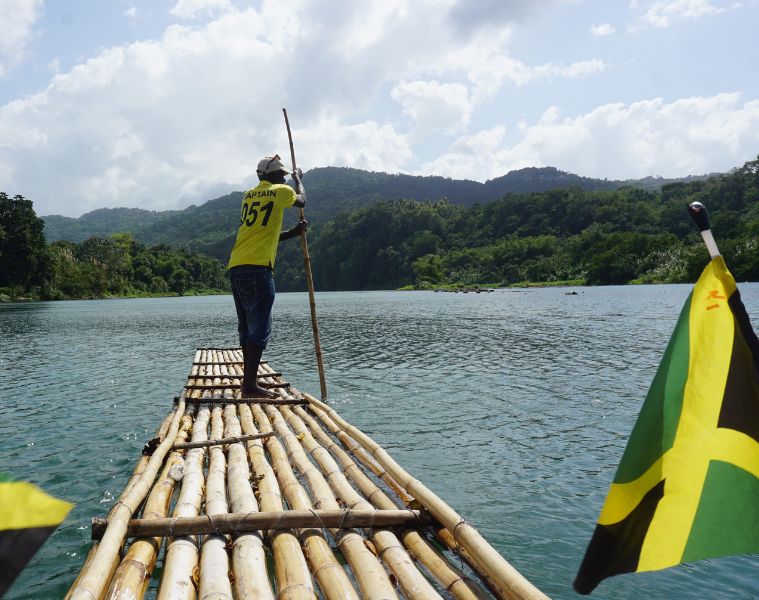 Rear view of a man bamboo rafting on the Martha Brae River in Jamaica, with a Jamaican flag on the raft, rowing toward scenic forested mountains.