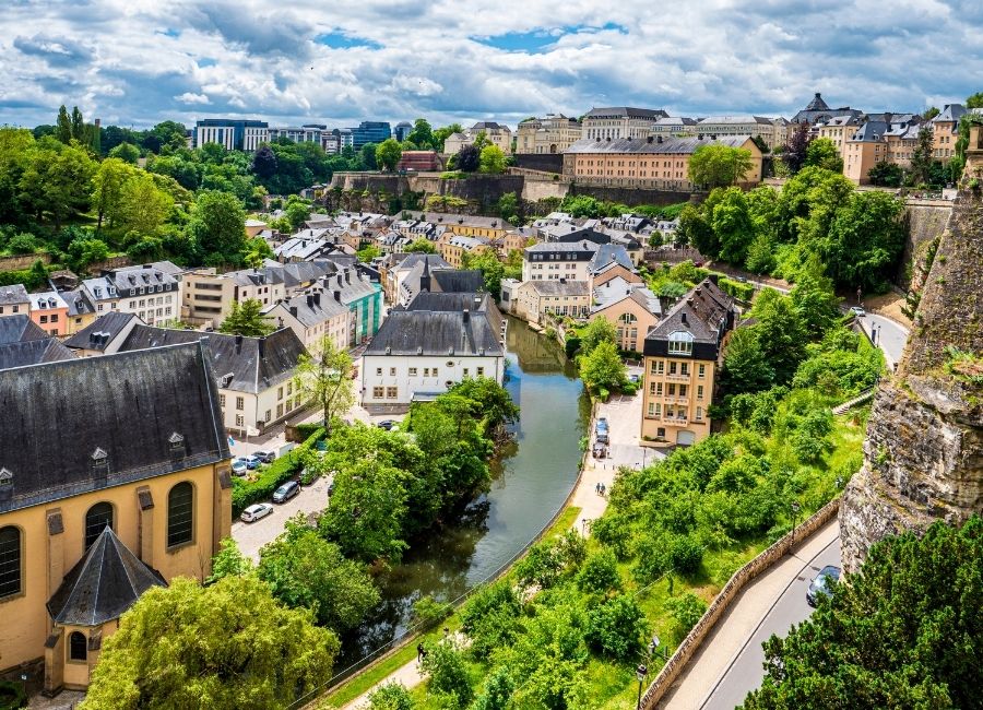 A scenic view of Luxembourg City with its old fortress walls, colorful houses along the Alzette River, and lush greenery