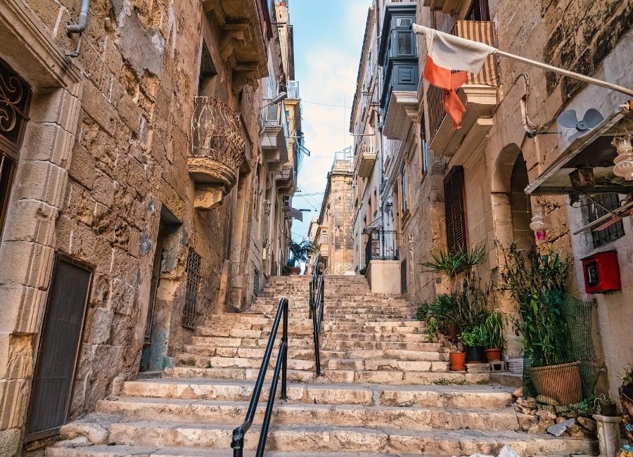 A narrow stone street with steep stairs in Valletta, Malta, surrounded by old limestone buildings and traditional Maltese balconies