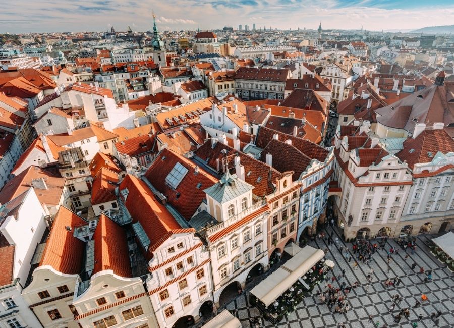 An aerial view of Prague's Old Town with red-tiled roofs, historic Baroque and Gothic architecture, with people congregating at the square below