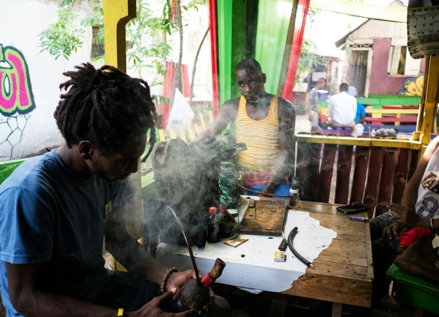 A group of Jamaican men sitting together in a colorful outdoor patio setting, surrounded by smoke, with one of the men holding a pipe