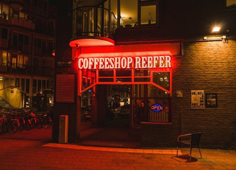 Front shot of an Amsterdam coffeeshop at night with a bright red neon sign reading 'Coffeeshop Reefer' above the entrance