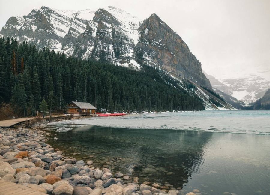 Snow-capped mountains and a pine forest surrounding Lake Louise in Banff National Park, Canada, with a wooden cabin and red canoes by the shore
