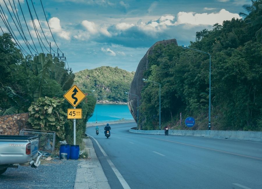 Curving coastal road in Koh Samui, Thailand, with motorbikes, green hills, and turquoise sea in the background