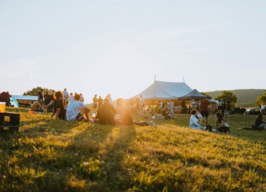 A group of people relaxing and socializing on a grassy field during sunset, with festival tents and a few trees in the background