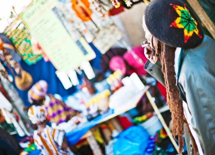 A Rastafarian man smoking at a vibrant street market with colourful handmade goods and Jamaican-inspired crafts