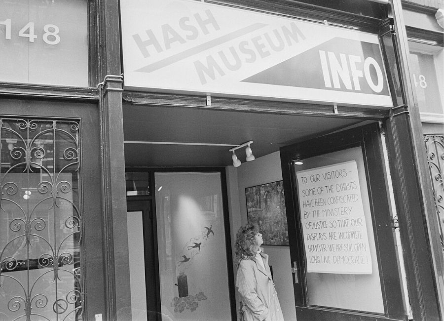 A 1987 black and white photograph of the entrance to the Hash Museum Info after a police raid, showing a sign with the text "HASH MUSEUM INFO" and a woman standing in the doorway. A notice posted in the window reads: "TO OUR VISITORS - SOME OF THE EXHIBITS HAVE BEEN CONFISCATED BY THE MINISTRY OF JUSTICE SO THAT OUR DISPLAYS ARE INCOMPLETE HOWEVER WE ARE STILL OPEN LONG LIVE DEMOCRACIE!"