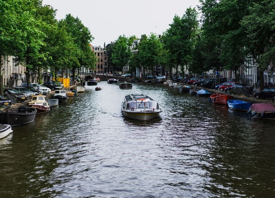 A canal in Amsterdam with a tour boat navigating the water, flanked by parked cars and tall green trees lining the banks under an overcast sky