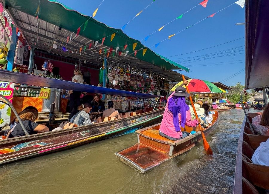 A view of a busy floating market in Bangkok, Thailand, with wooden longtail boats carrying vendors and tourists along a narrow waterway, surrounded by stalls selling goods