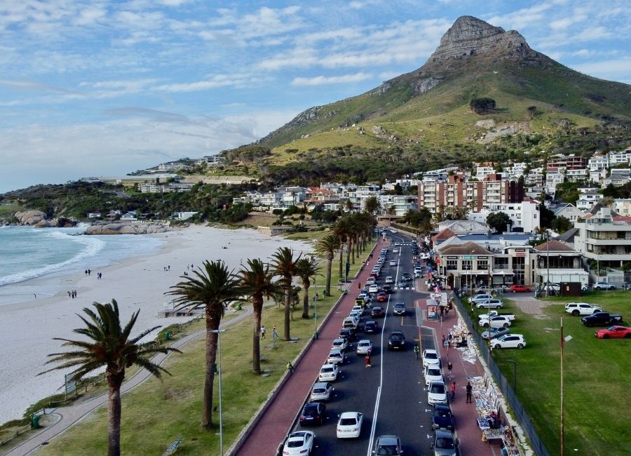 A panoramic view of Camps Bay strip in Cape Town, South Africa, featuring a busy coastal road lined with palm trees, a sandy beach, and residential buildings leading up to Lion's Head mountain in the background