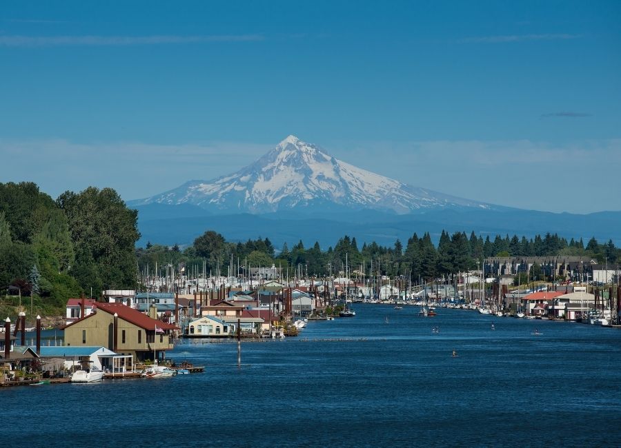 A view of the snow-capped Mount Hood towering over a body of water lined with houseboats, marinas, and greenery in the foreground under a blue sky